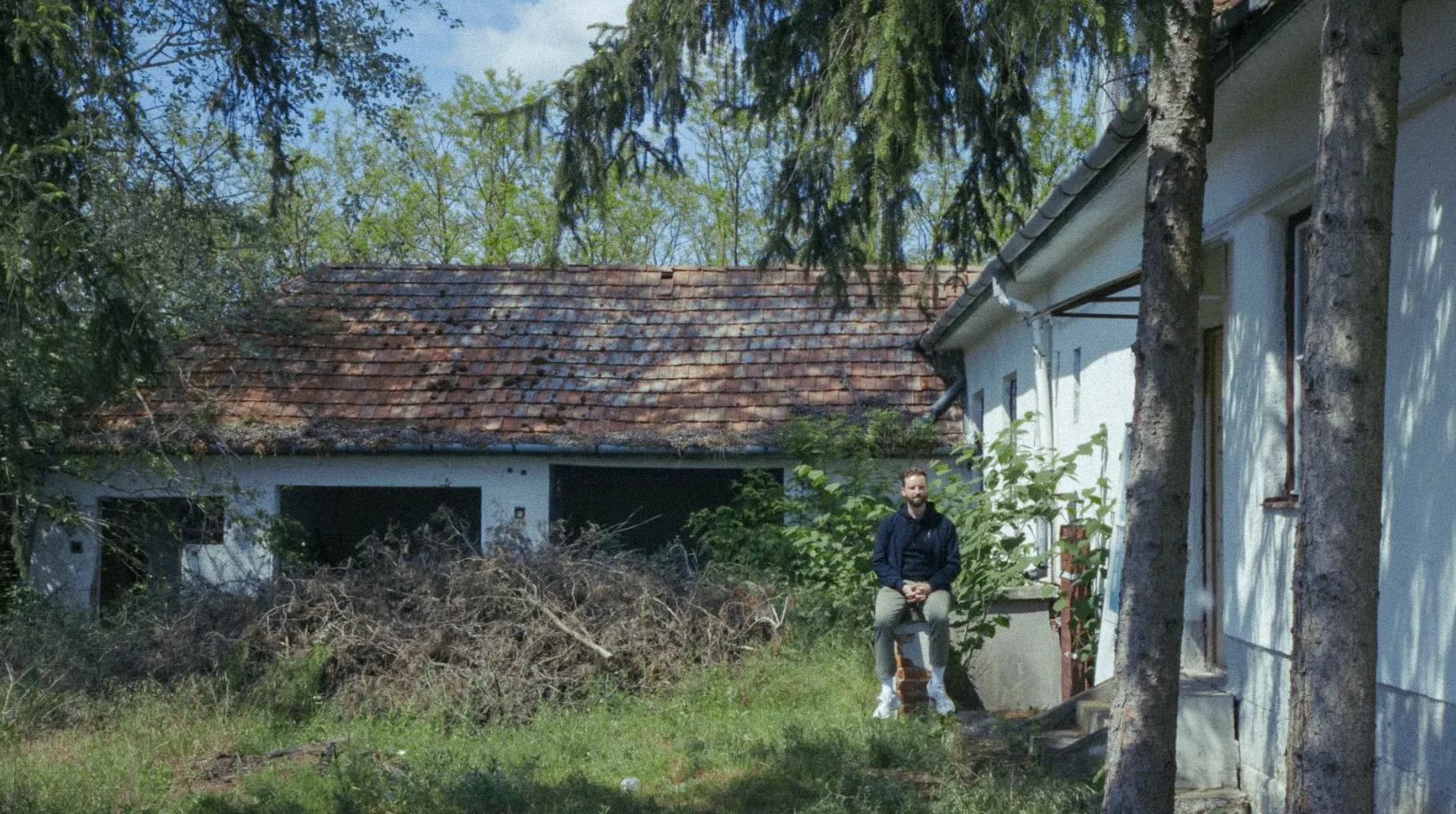 Gulyás Márton egy széken ül a felújításra váró penészleki ház előtt, benőtt kerttel körülvéve.
